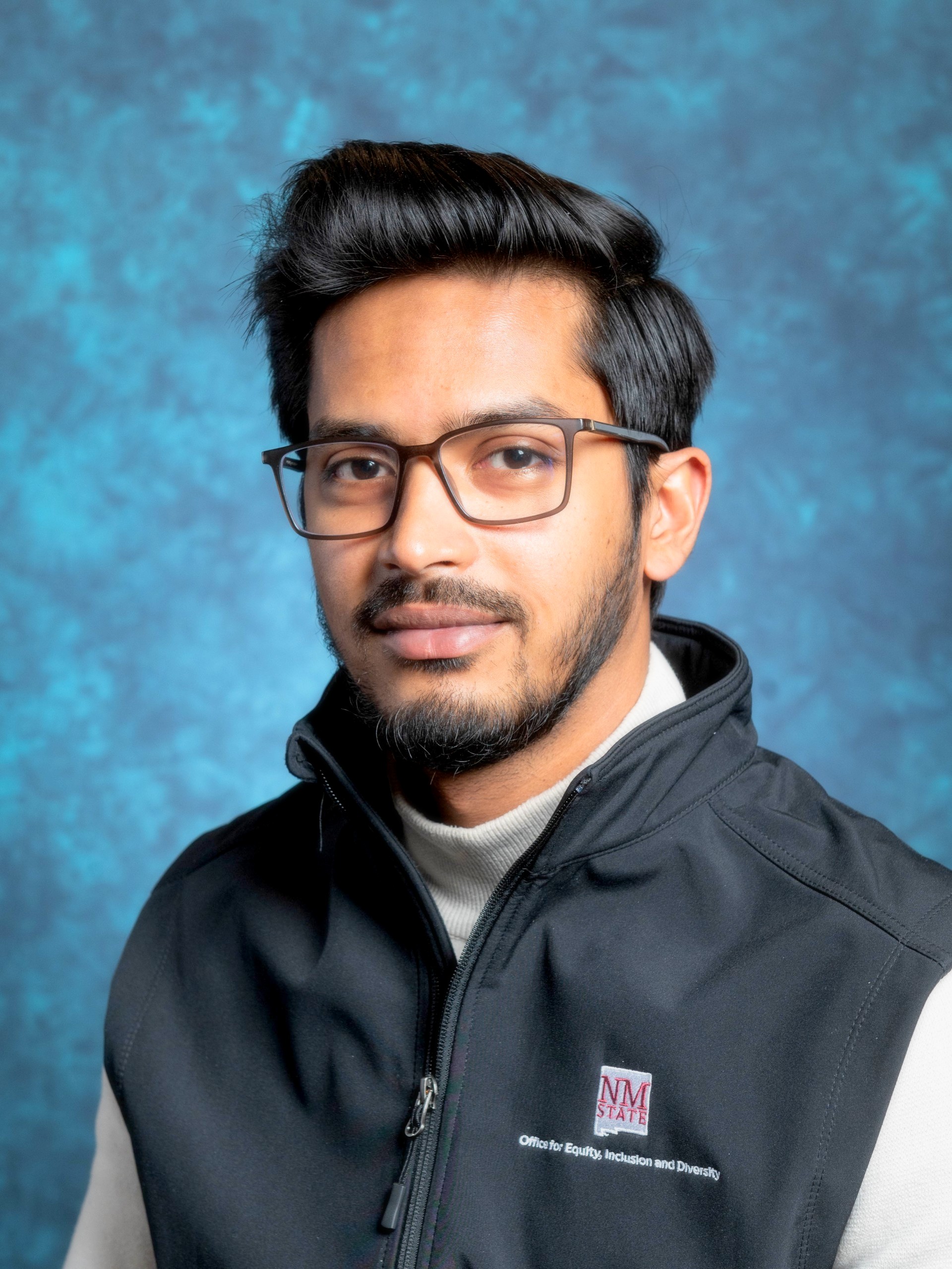 An image of Sarhan Osman Bhuiya, the Graduate Assistant for EID. The image is a head-and-shoulders portrait of a Sarhan against a textured blue background. The person has short, dark hair with a slight wave and a well-groomed beard and mustache. He is wearing rectangular glasses with brown frames and are gazing directly at the camera with a neutral expression. He is dressed in a black zip-up vest over a light gray turtleneck. The vest has an emblem on the left chest area with the text "NM STATE" in maroon and a small patch of additional text in white below it.